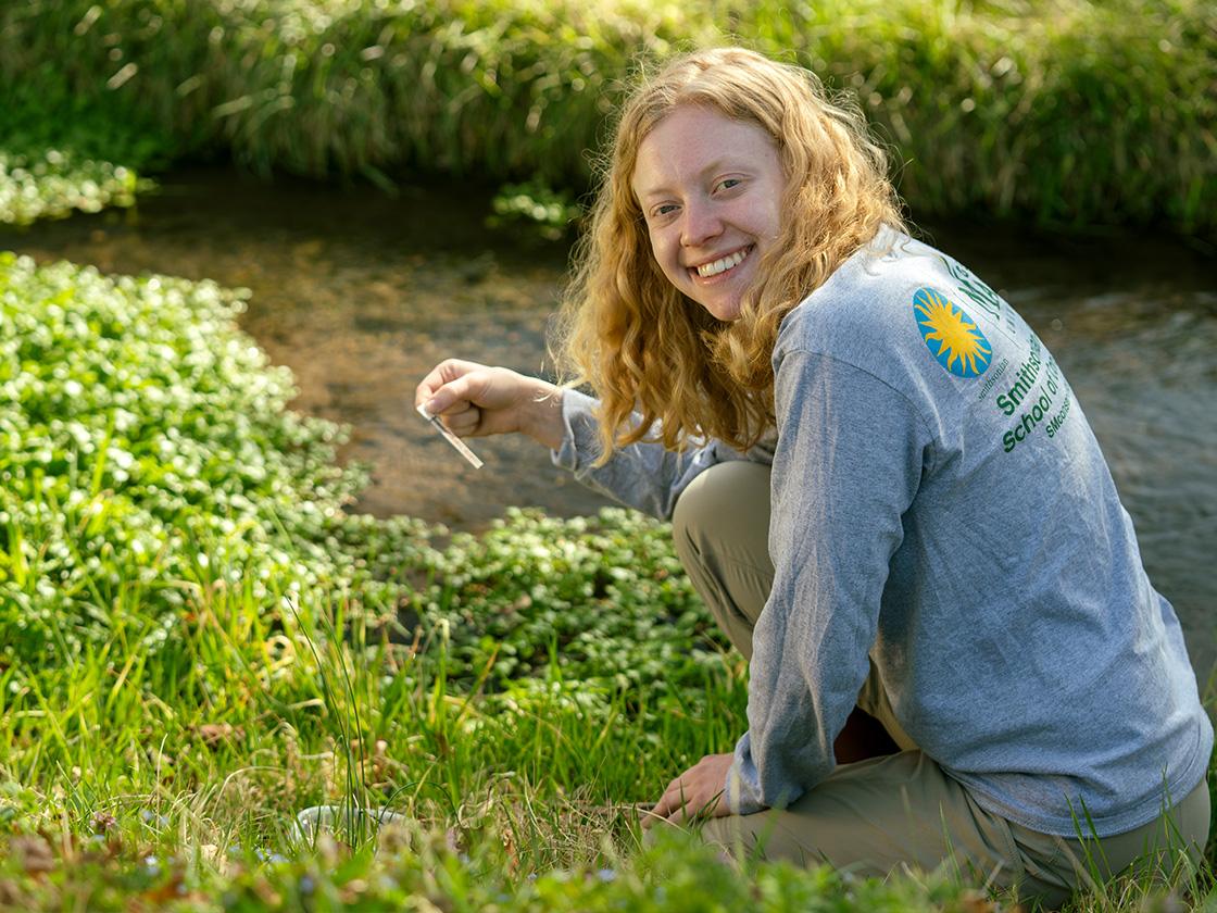 Smiling student sitting by a river while taking samples and wearing a Smithsonian t-shirt