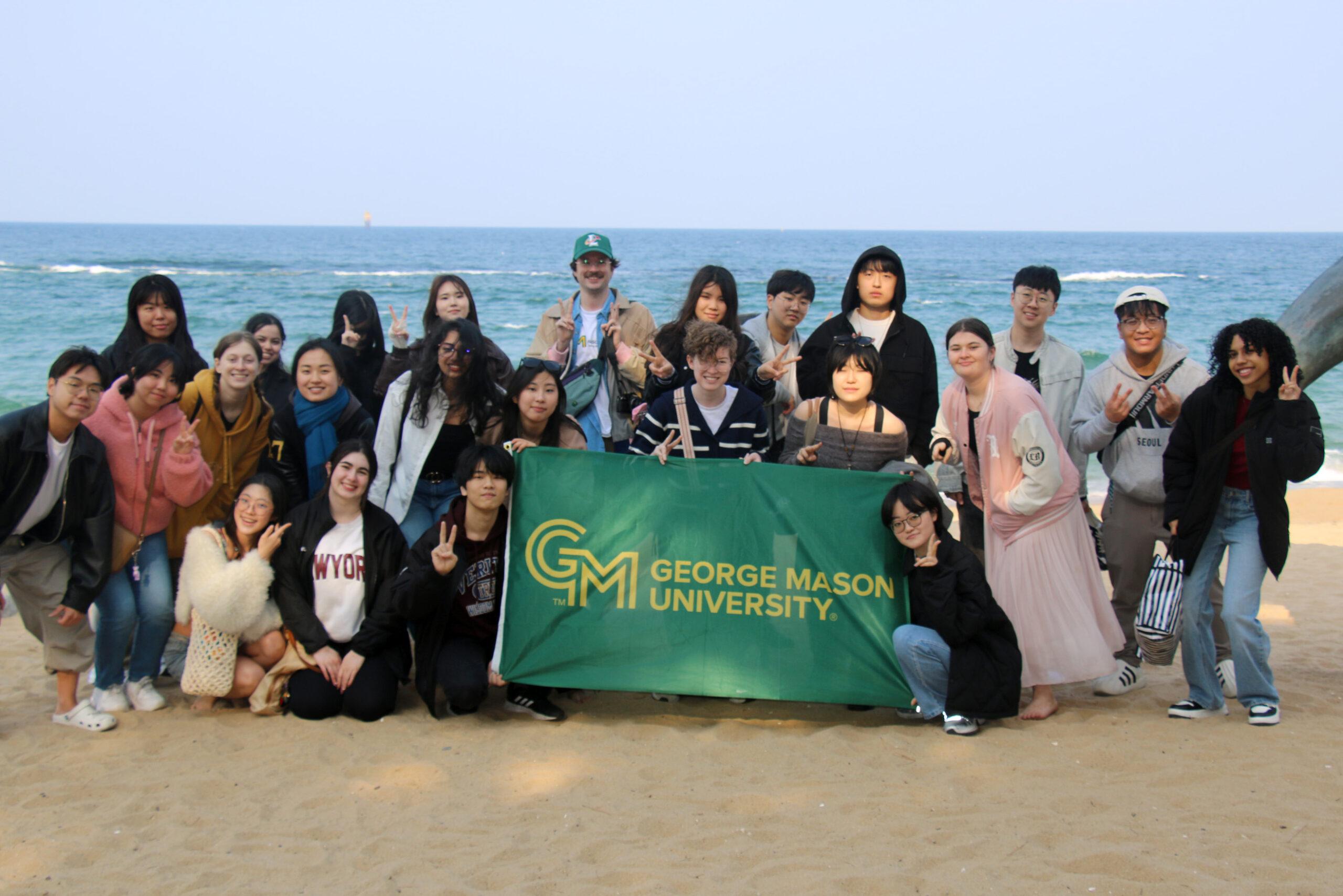 A group of students on the beach and holding a green sign with George Mason's logo