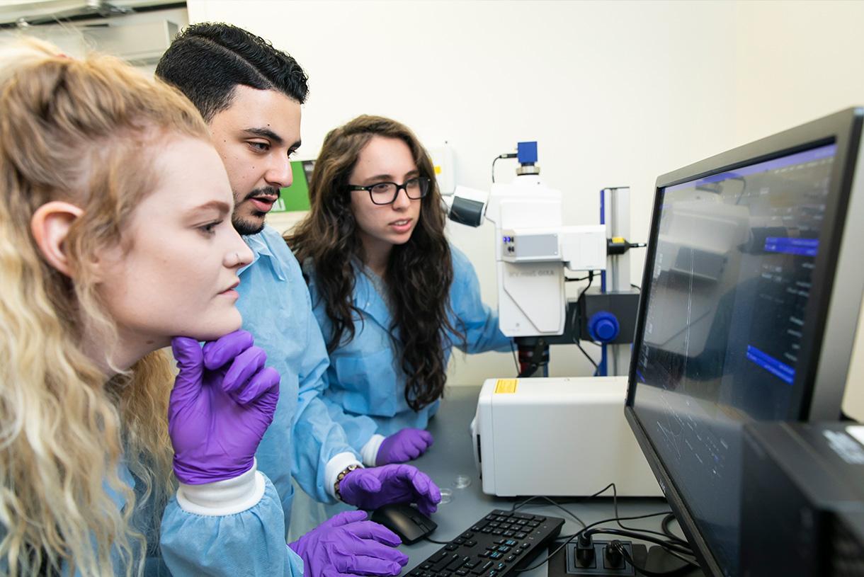 Three students wearing lab coats and protective gloves and looking at a desktop screen