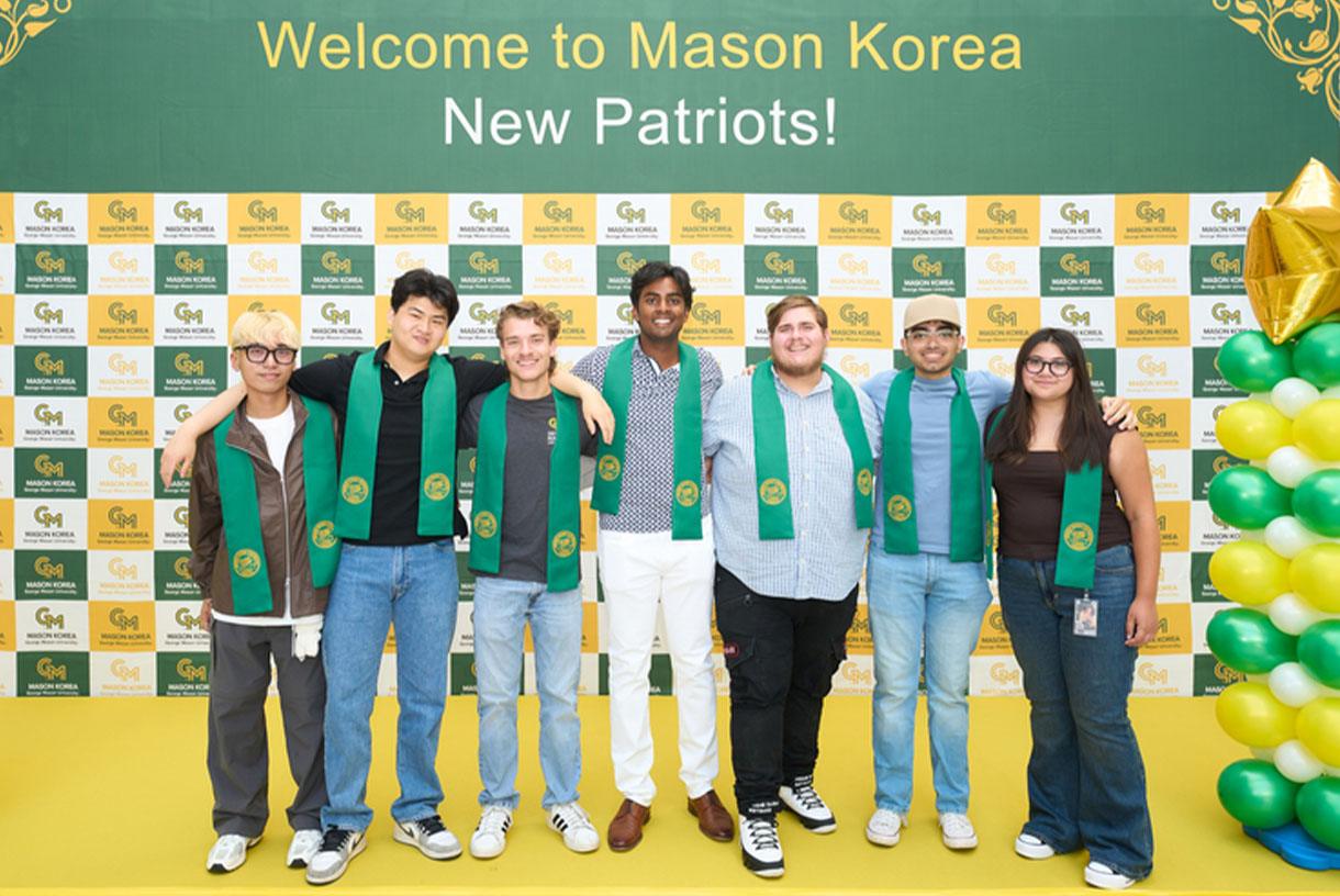Students wearing green sashes and standing in front of a green and yellow sign that says Welcome to Mason Korea