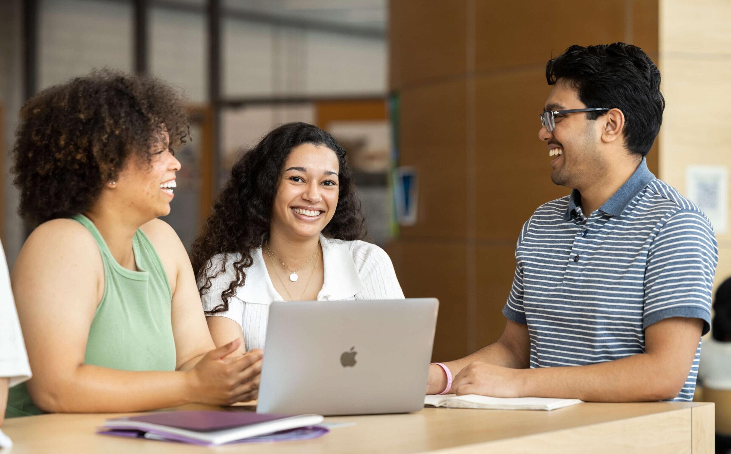 Three smiling students working together at a table and laptop