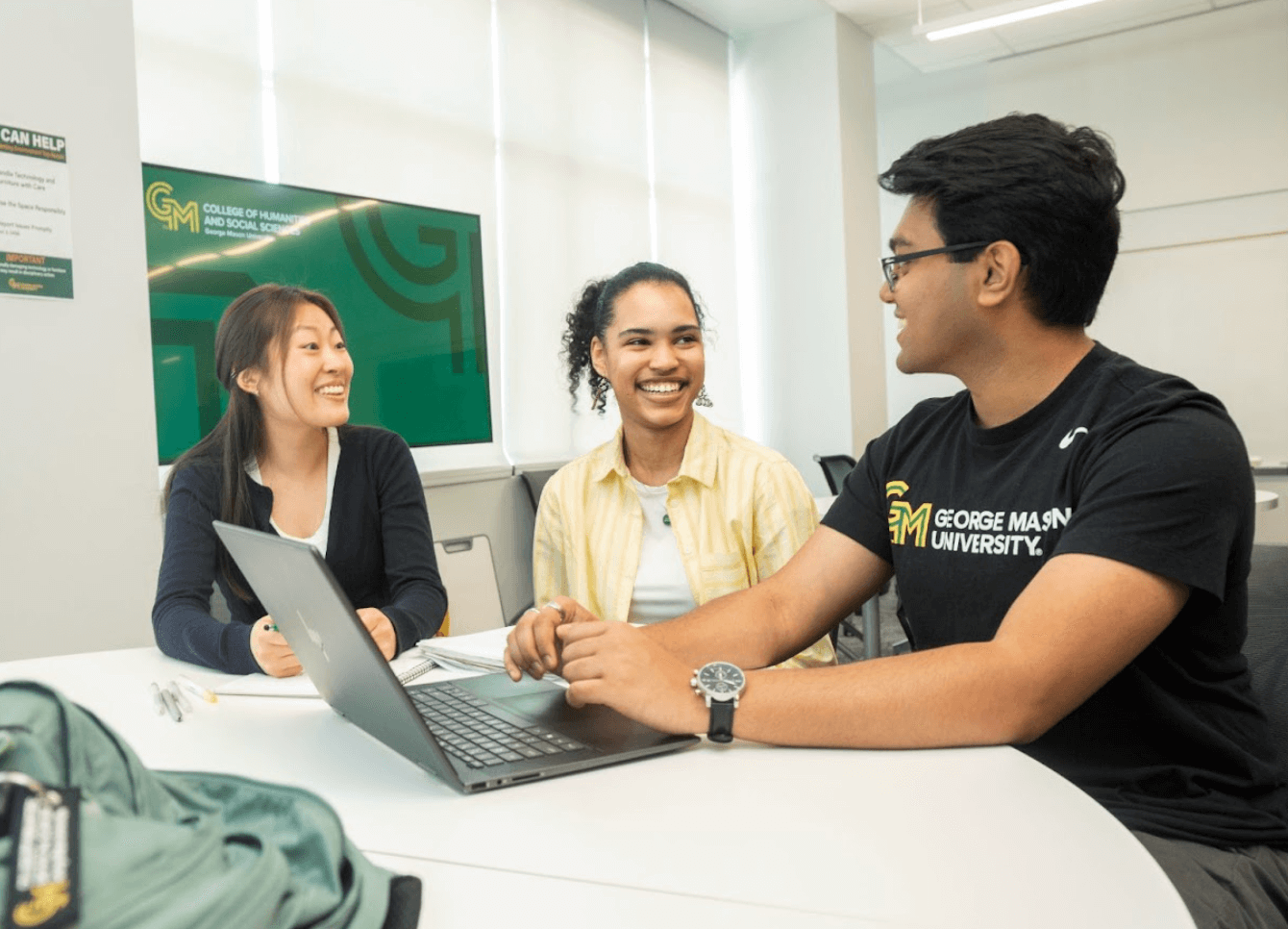 Three smiling students sitting at a table and working at a laptop
