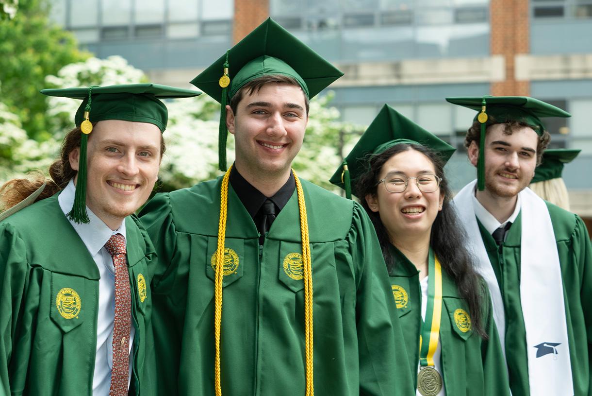 Four smiling students wearing green commencement cap and gown