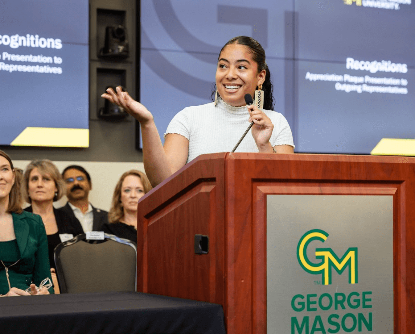 Smiling student holding a microphone and standing behind podium with George Mason signage