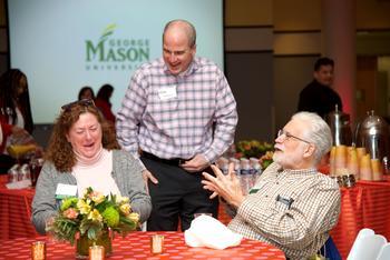A group of three faculty and staff together at the Faculty and Staff Donor Breakfast event on the Fairfax Campus. They are talking and laughing. The table is set with colorful linens, votives, and a flower arrangement. The presentation screen in the background has a George Mason University logo displayed.