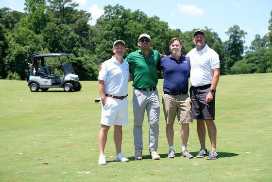 Four golfers pose on the green with a golf cart in the background.