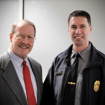 A man in a red tie stands next to a man in a jacket with a badge.