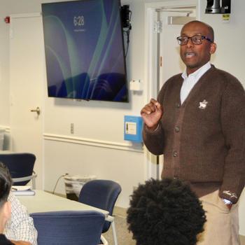 A man in a brown sweater stands in front of a classroom.
