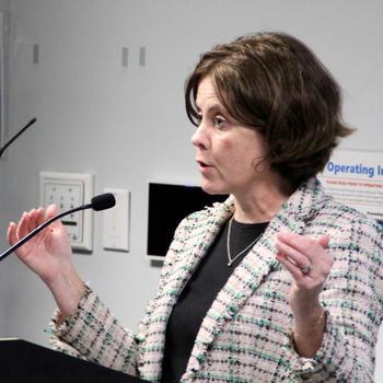 A woman in a light jacket and is speaking into a microphone.