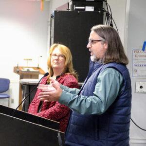 A woman in a red sweater and a man in a blue vest engage an audience.