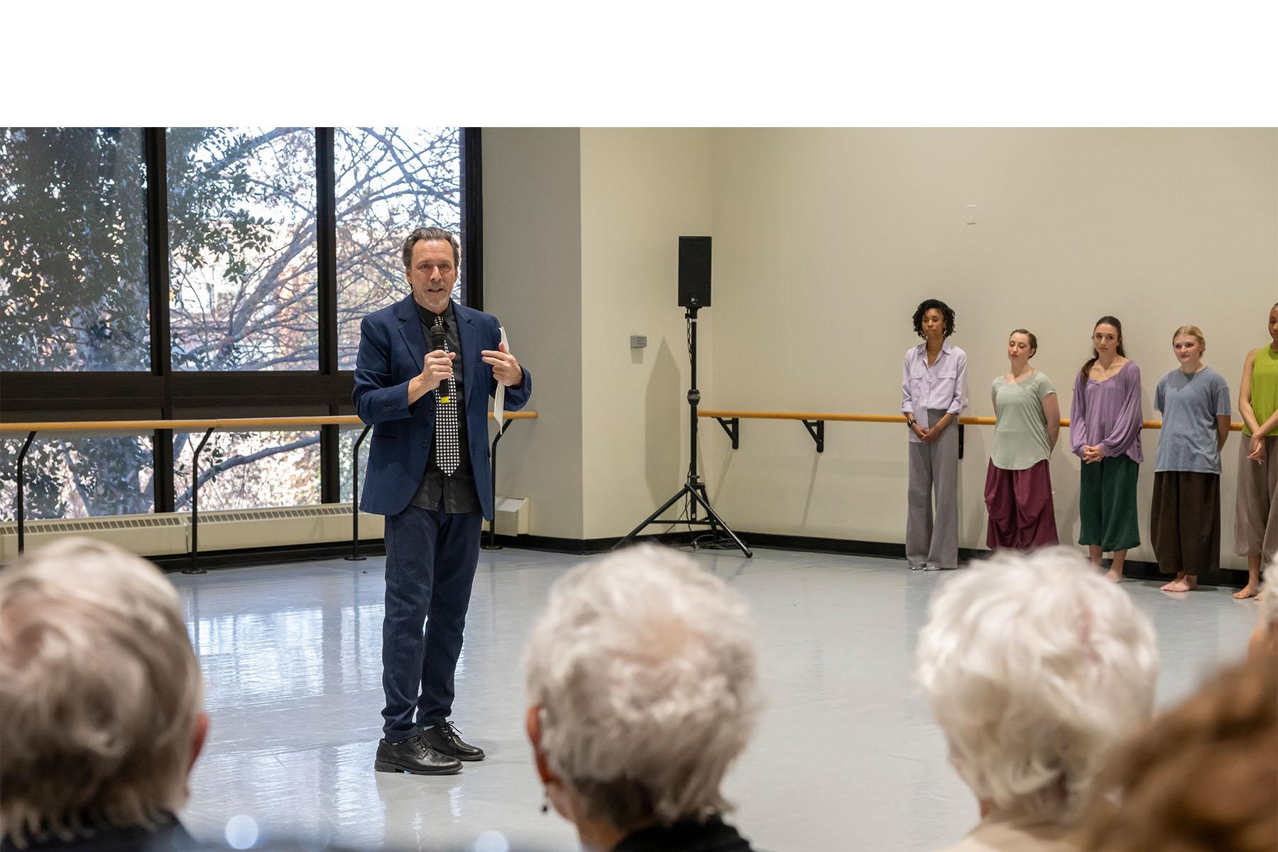 A speaker stands in a dance studio with dancers lined up in the background.