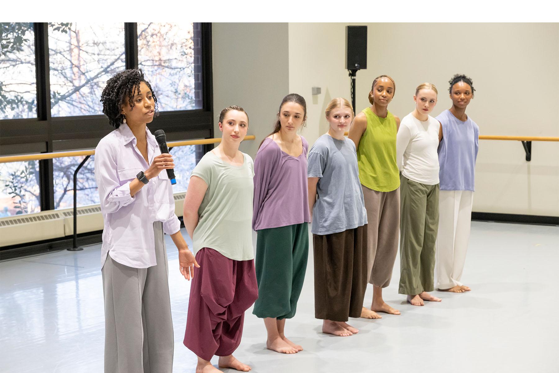 A speaker stands in a dance studio with six dancers beside them.